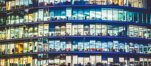 exterior shot of an office building windows lit up at night, with workers silhouetted inside