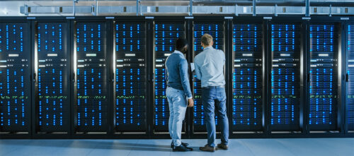 A Black engineer and a white engineer look at a rack of servers in a data center.