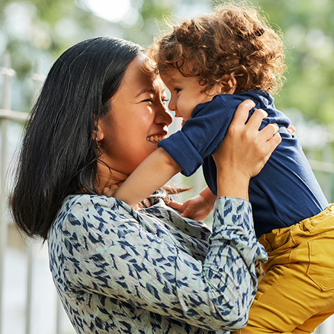 An East Asian mother holds her young toddler. They are both looking at each other and smiling.