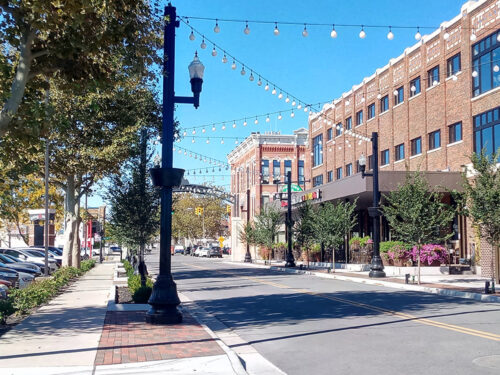 An image of a downtown streetscape, with a four-story brick building fronting a two-lane street adorned with ornamental pedestrian lighting, street trees, and a sidewalk cafe.