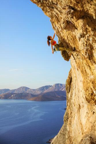 Female rock climber on challenging route, looking up at cliff, view of coast below