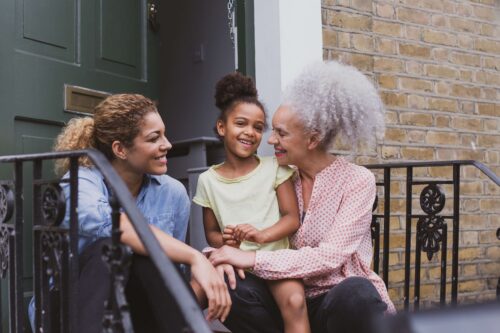An intergenerational Black family - a mother, child, and grandmother- sit on front steps to a home.