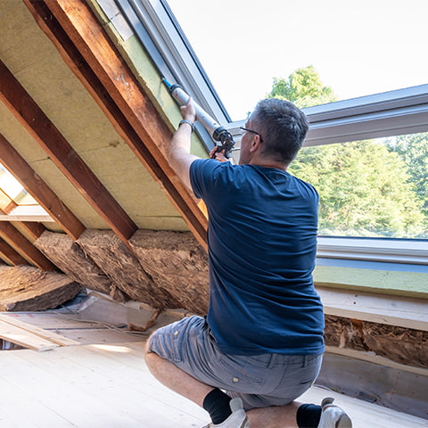 A middle-aged white craftsman caulks an attic window.