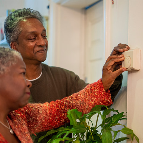 An elderly mixed-race couple adjusts a thermostat in their home