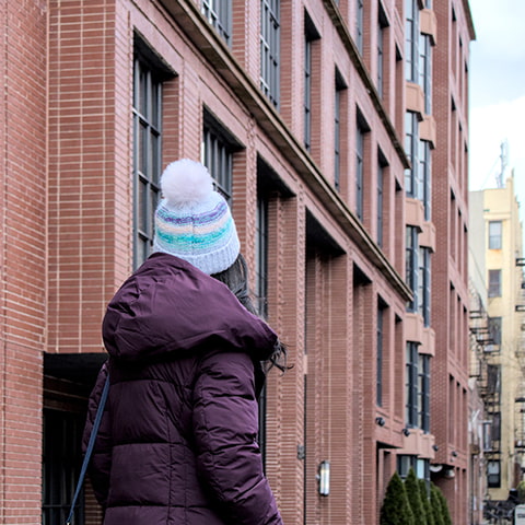A woman in a winter jacket and a beanie looks up at dense New York City apartment buildings.