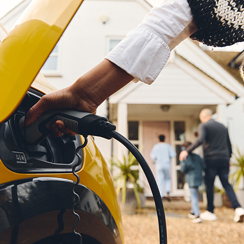 A Black woman inserts a charger into a yellow electric car. In the background, her mixed-race family enters their home