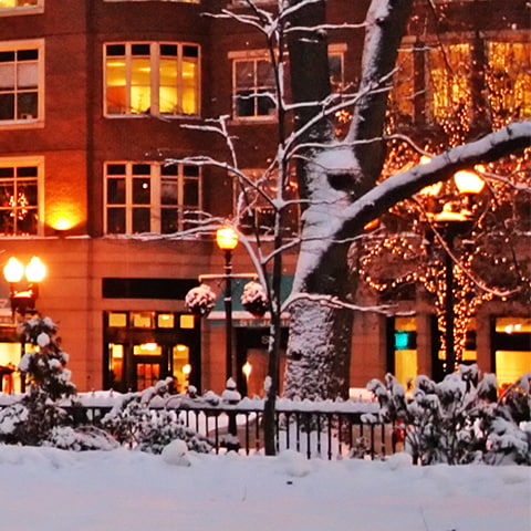A winter evening in Boston, Massachusetts. Snow covers the ground; apartment lights and streetlights are on,.