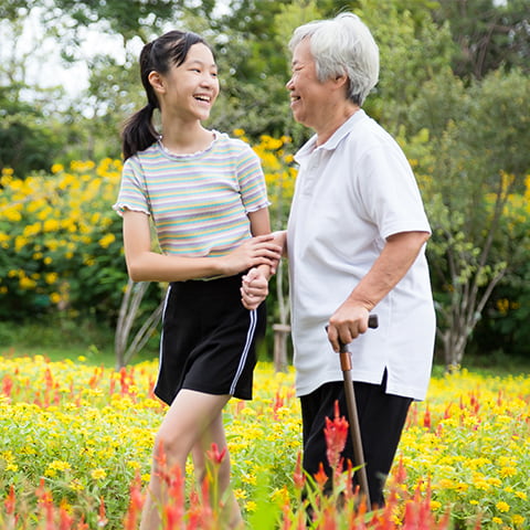 An East Asian granddaughter helps her elderly grandmother walk through a field of blooming flowers