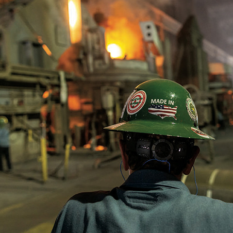 A view from behind an industrial worker looking at a furnace. Their dark green helmet features a “made in the USA” sticker.