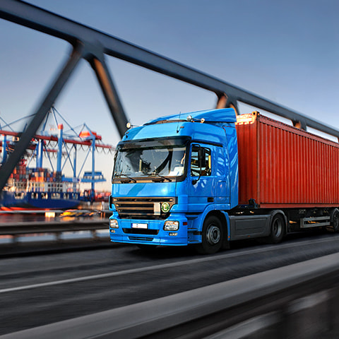A semi truck drives across a bridge. In the background, cranes unload shipping containers from a container ship at a busy port.